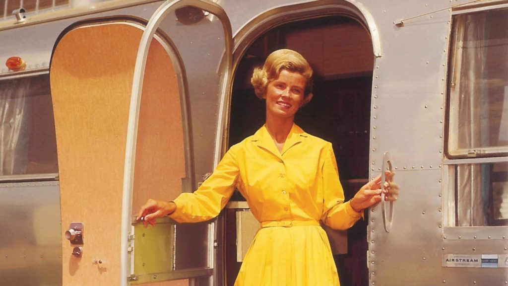 Una donna con un vestito giallo anni cinquanta che guarda fuori dalla porta di una Airstream. Sorride, è soddisfatta e felice.
