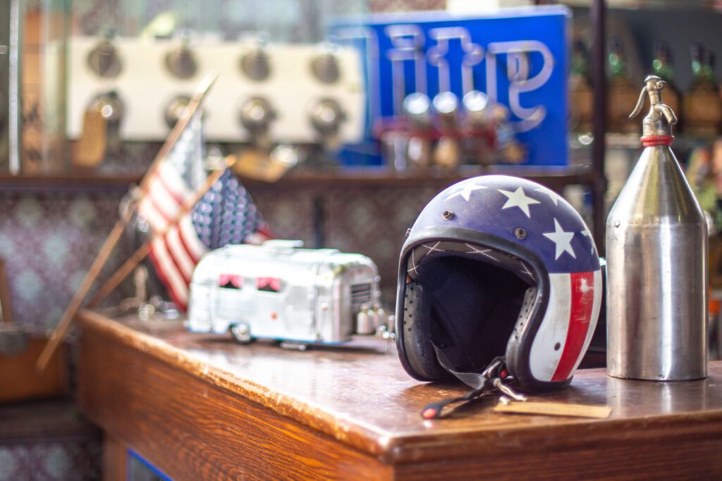 dettaglio di un tavolo di legno con sopra un casco da moto decorato a stelle e strisce, una miniatura di una Airstream e delle bandiere americane sullo sfondo 