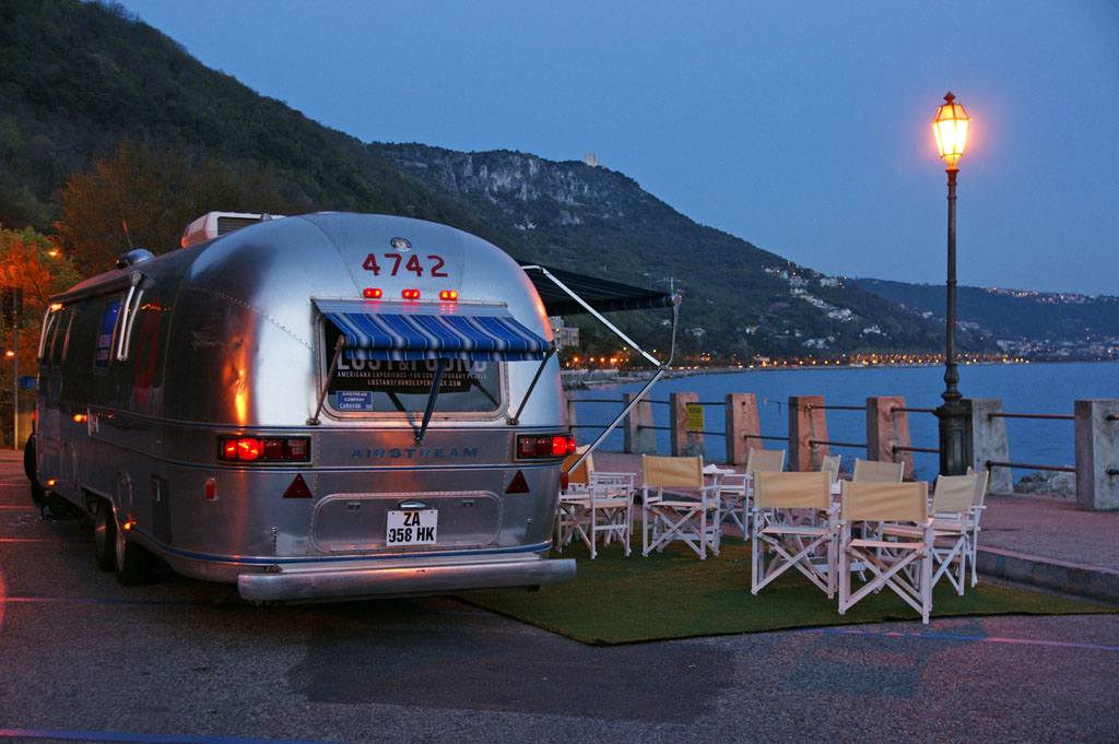 Airstream Lounge in una località marittima al tramonto, con dei lampioni a mostrare la scena. alcune sedie di fronte al mezzo