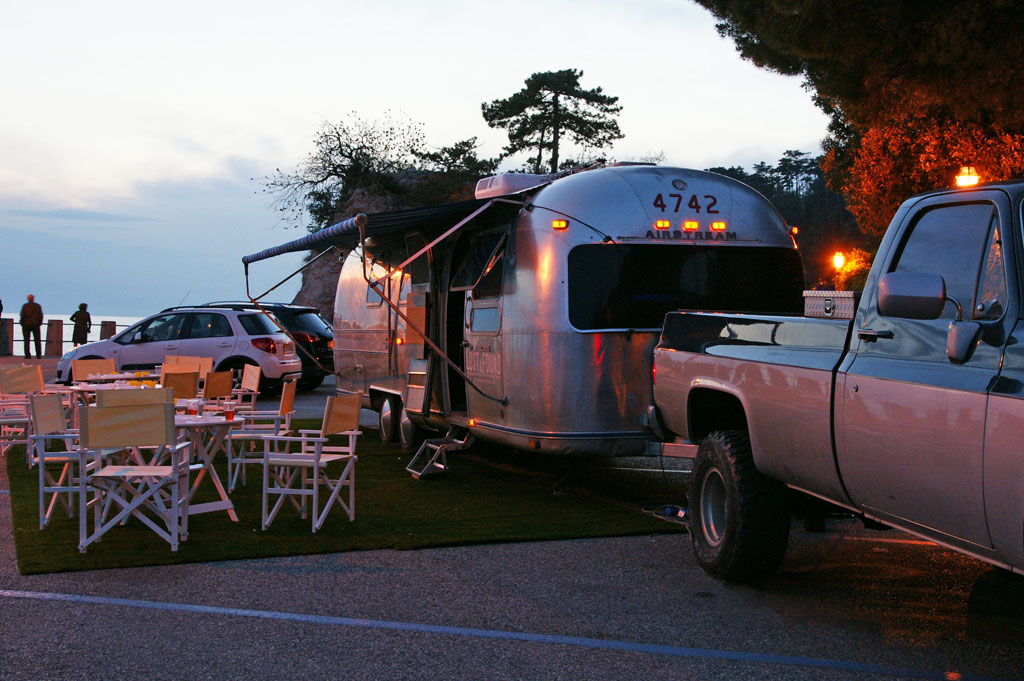Airstream Lounge al tramonto, con luci calde sullo sfondo e una tenda aperta. Tavole e sedie di fronte alla porticina che concede l'accesso al mezzo, macchine sul background.